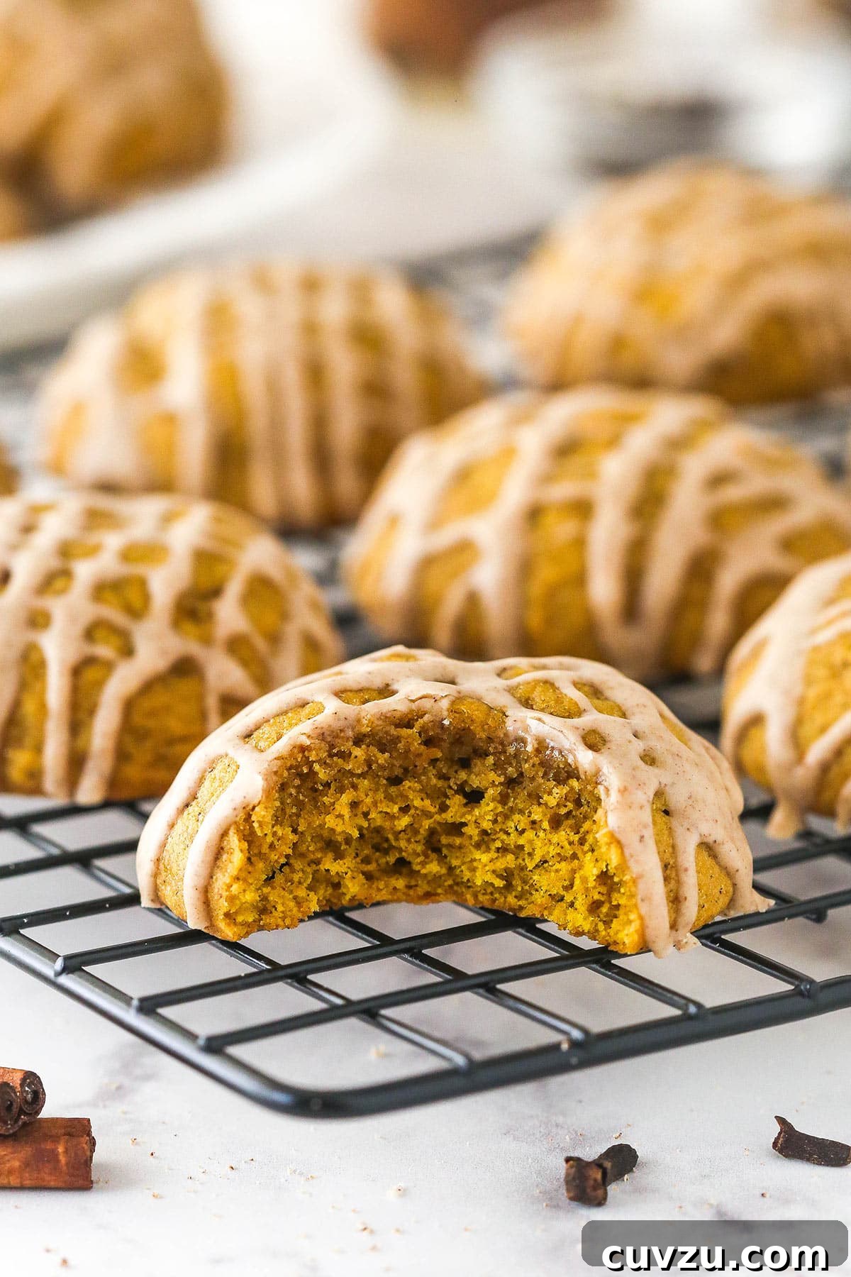 Pillowy Pumpkin Delights 4 Pumpkin cookies with cinnamon glaze on a cooling rack, one cookie has a bite taken out of it.