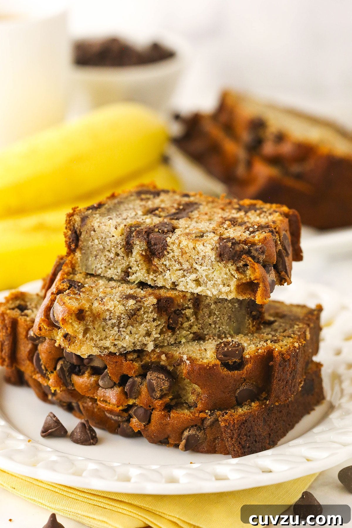 This easy Chocolate Chip Banana Bread has the moistest crumb & the softest, fluffiest texture. You'll never follow a different recipe again! Slices of chocolate chip banana bread stacked onto a plate with two bananas in the background