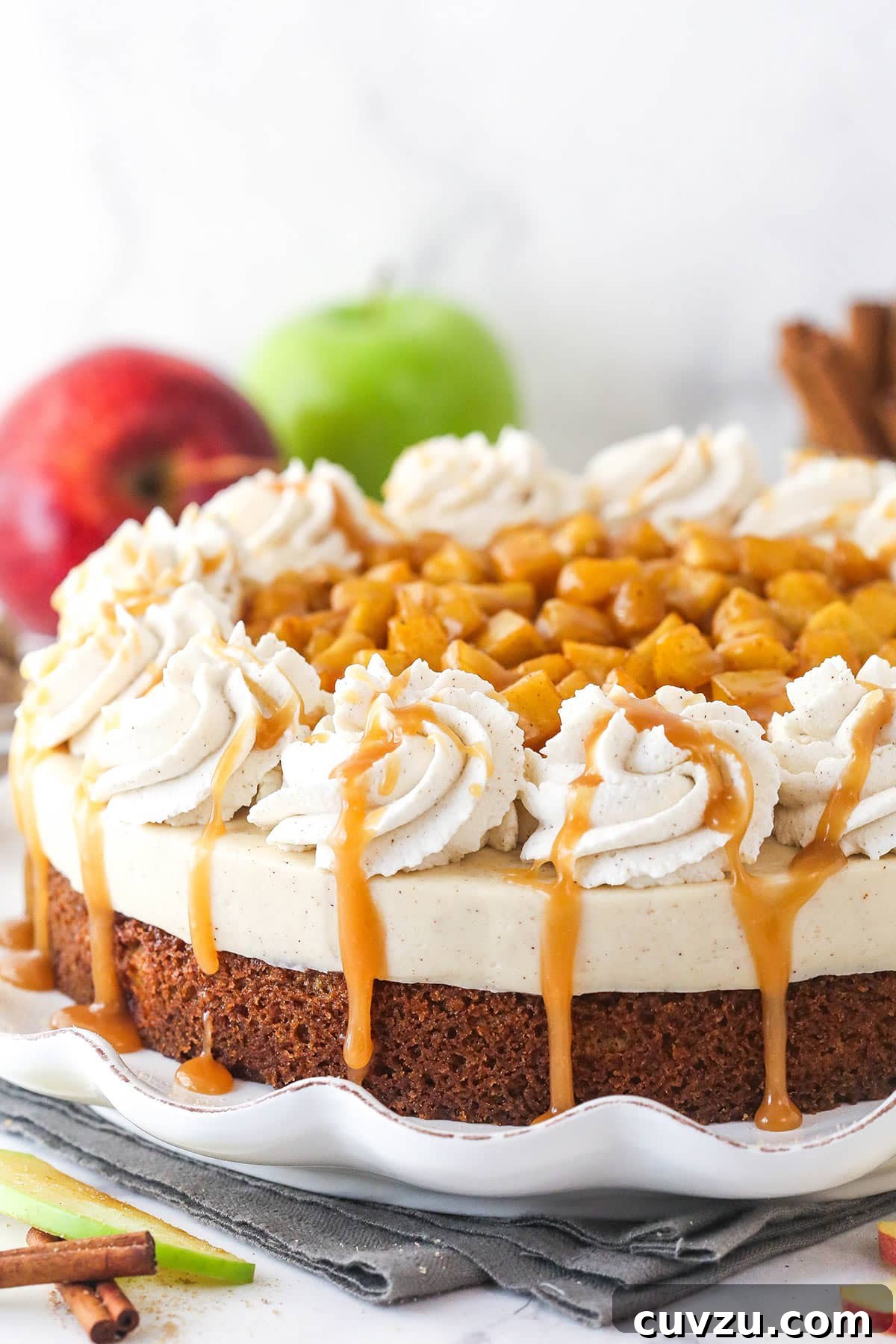 Decadent Caramel Apple Blondie Cheesecake 12 A beautifully decorated Caramel Apple Blondie Cheesecake on a white ruffled cake plate, topped with piped whipped cream, cinnamon apples, and caramel drizzle.