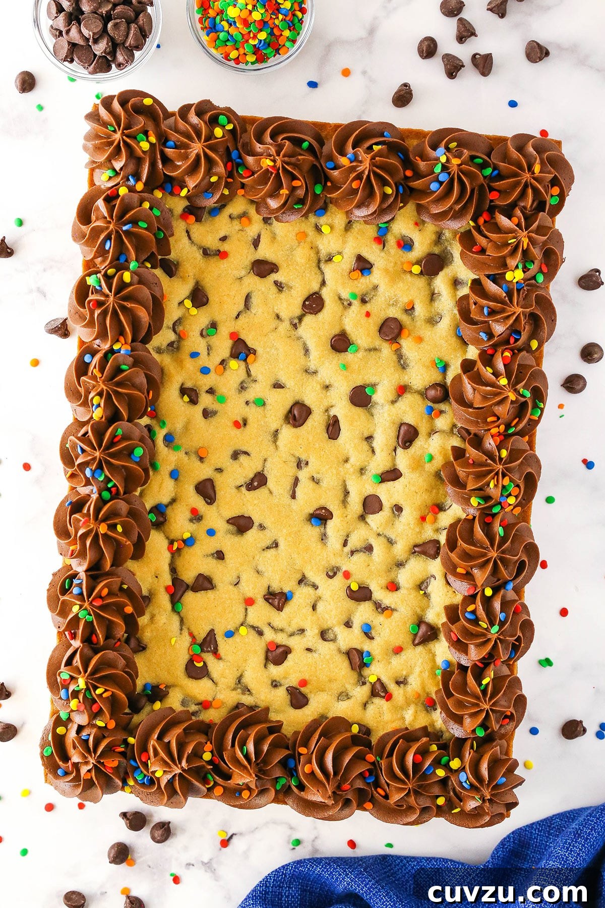 This easy Sheet Pan Cookie Cake features loads of chocolate chips and a rim of creamy chocolate frosting. It's perfect for feeding a crowd! A sheet pan cookie cake on a marble surface with chocolate chips surrounding it