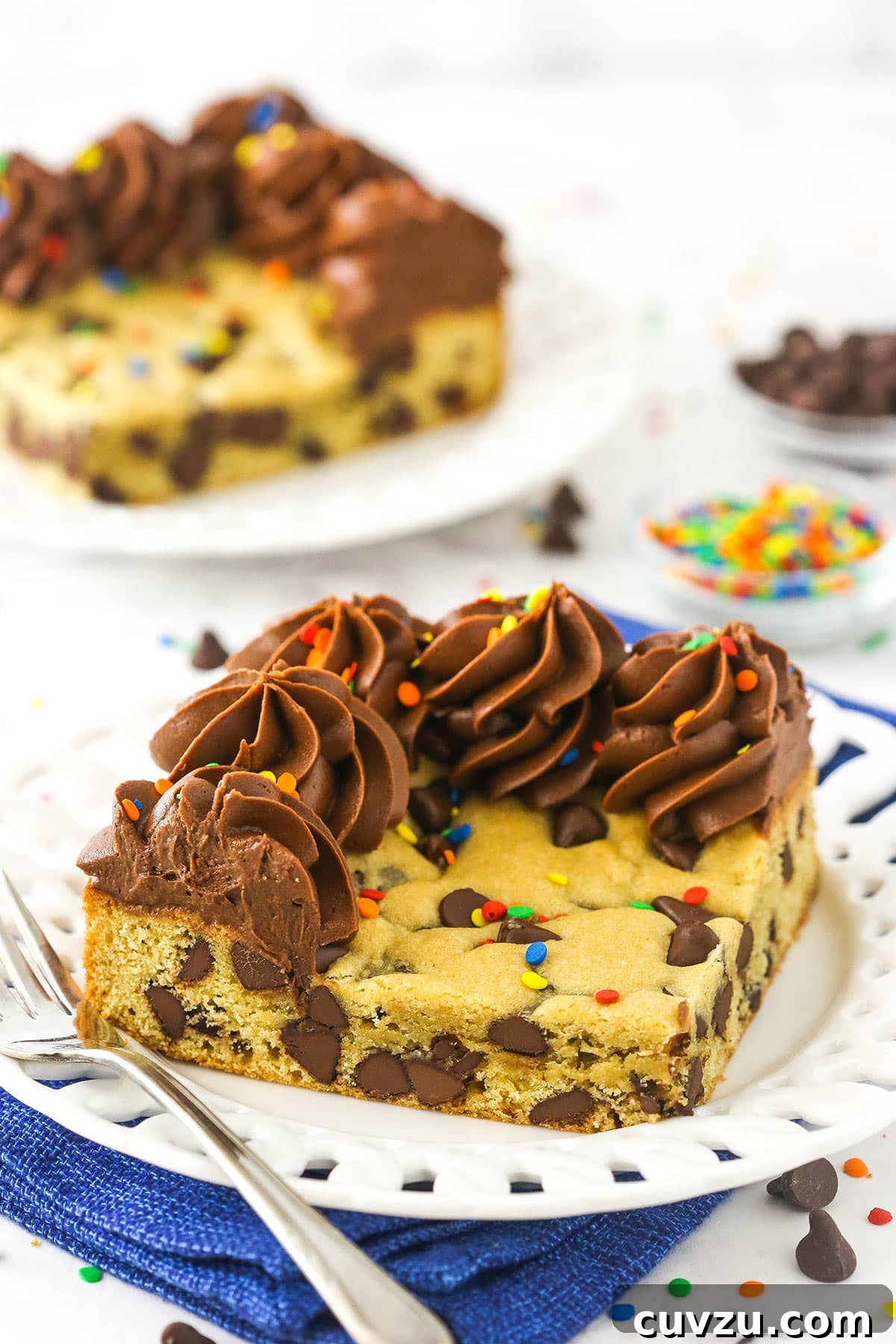This easy Sheet Pan Cookie Cake features loads of chocolate chips and a rim of creamy chocolate frosting. It's perfect for feeding a crowd! A piece of chocolate chip cookie cake topped with chocolate buttercream with a second slice behind it