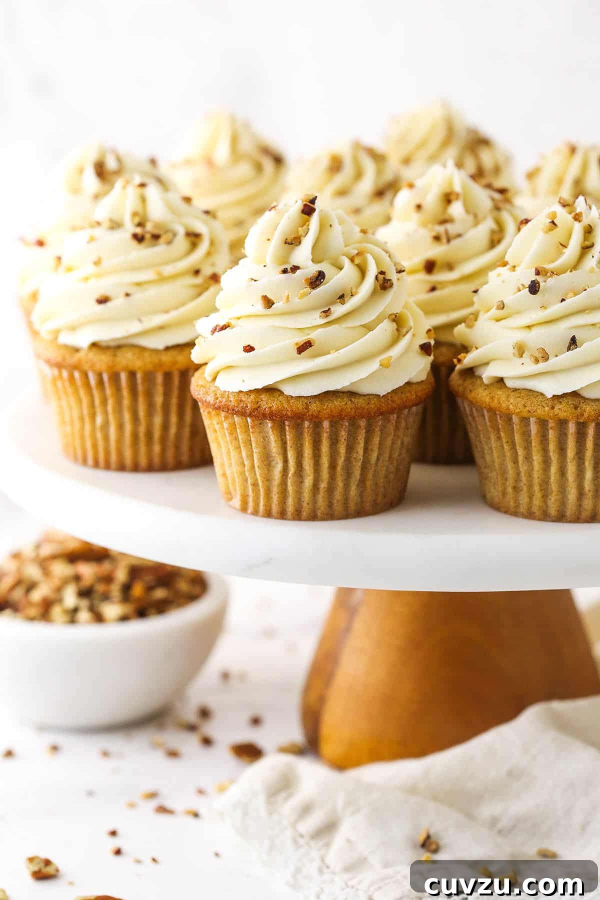 Soft brown sugar cupcakes are stuffed with gooey pecan pie filling & topped with creamy pecan pie frosting in this decadent homemade dessert. A beautiful display of freshly baked Pecan Pie Cupcakes on a rustic cake stand, with a bowl of toasted and chopped pecans beside them, ready for garnishing.