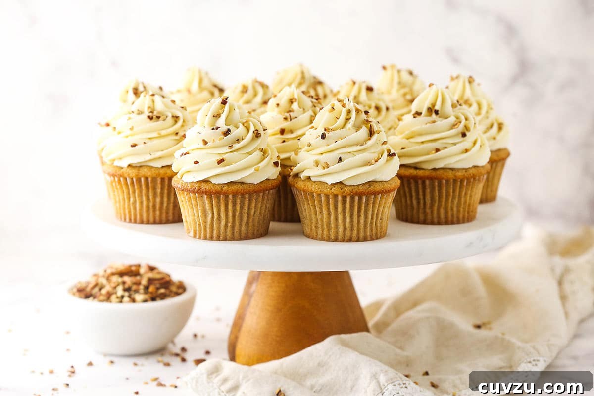 Soft brown sugar cupcakes are stuffed with gooey pecan pie filling & topped with creamy pecan pie frosting in this decadent homemade dessert. A charming scene of Pecan Pie Cupcakes arranged on a cake stand atop a cream-colored dishtowel on a countertop, ready for serving.