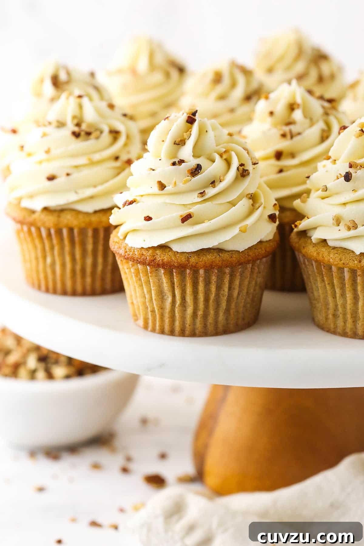 Soft brown sugar cupcakes are stuffed with gooey pecan pie filling & topped with creamy pecan pie frosting in this decadent homemade dessert. A full batch of beautifully frosted Pecan Pie Cupcakes arranged on a plastic cake stand with a wooden base, showcasing their tempting appeal.