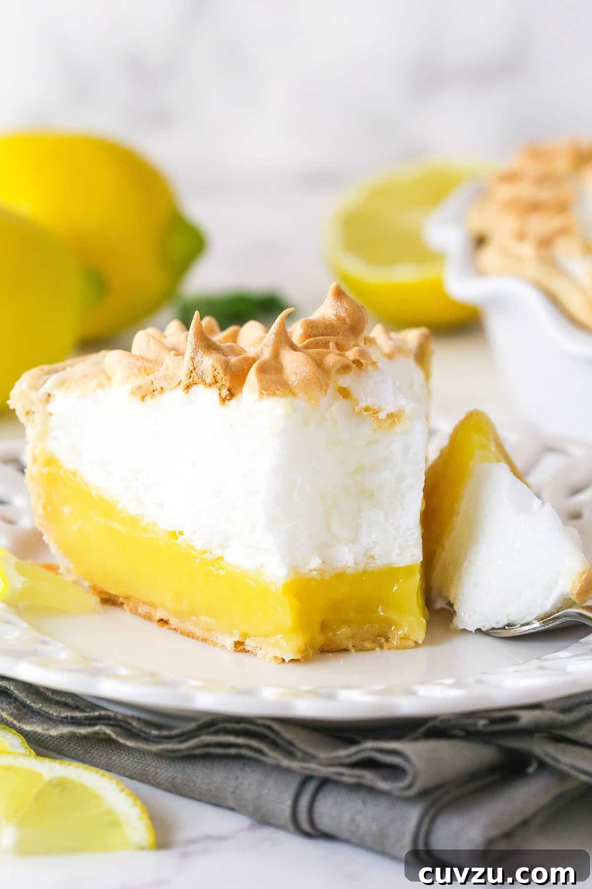 This classic lemon meringue pie features a tart lemon filling in a buttery crust topped with a light but stable meringue. Front view of a slice of lemon meringue pie on a plate with a bite on a fork